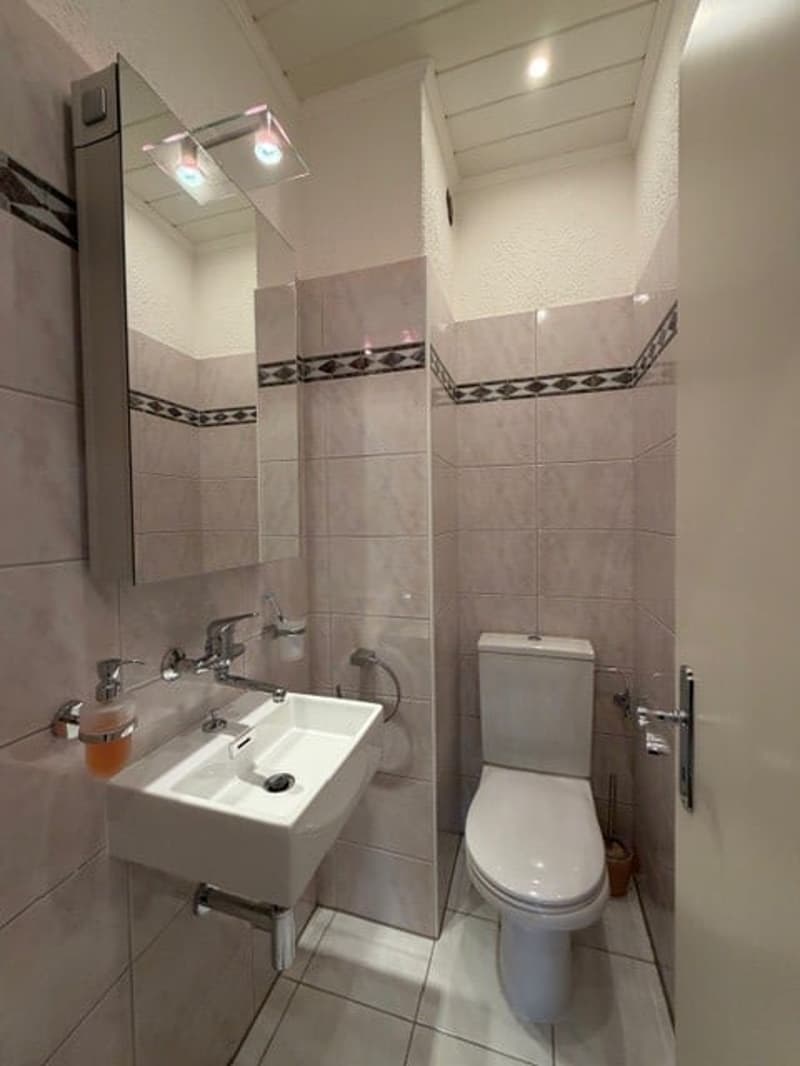 Bathroom with white tiles, a toilet, a sink, a mirror, and a soap dispenser.
