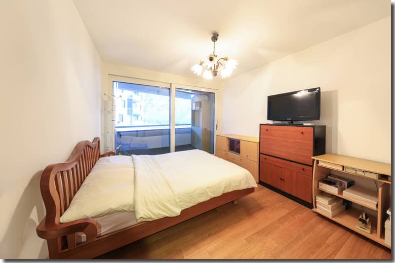 Wooden bed, wooden cabinet with TV, open balcony with balcony table and chairs, sliding glass door, lamp, wooden floor, white walls