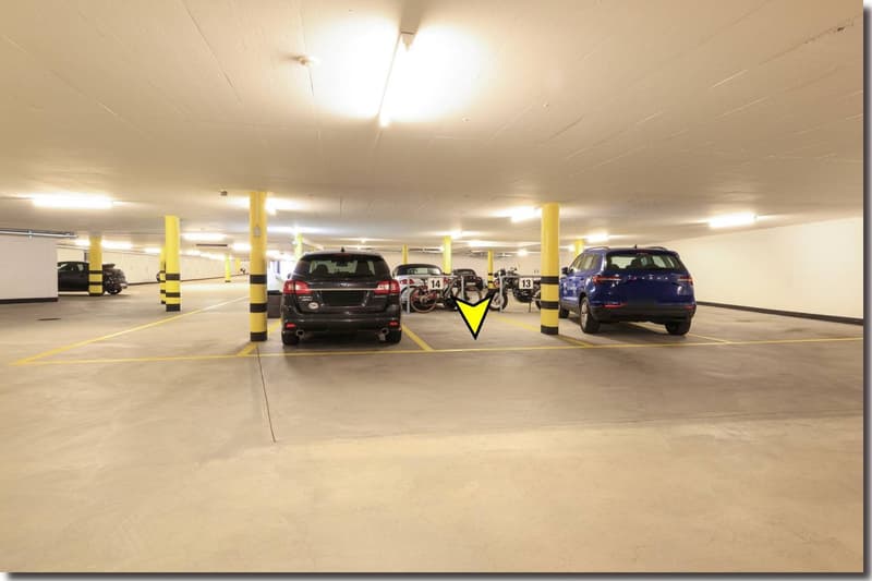 A covered parking lot with multiple cars parked, concrete floor, yellow lines marking parking spaces, white ceiling with lights, yellow arrows on the floor indicating parking spaces
