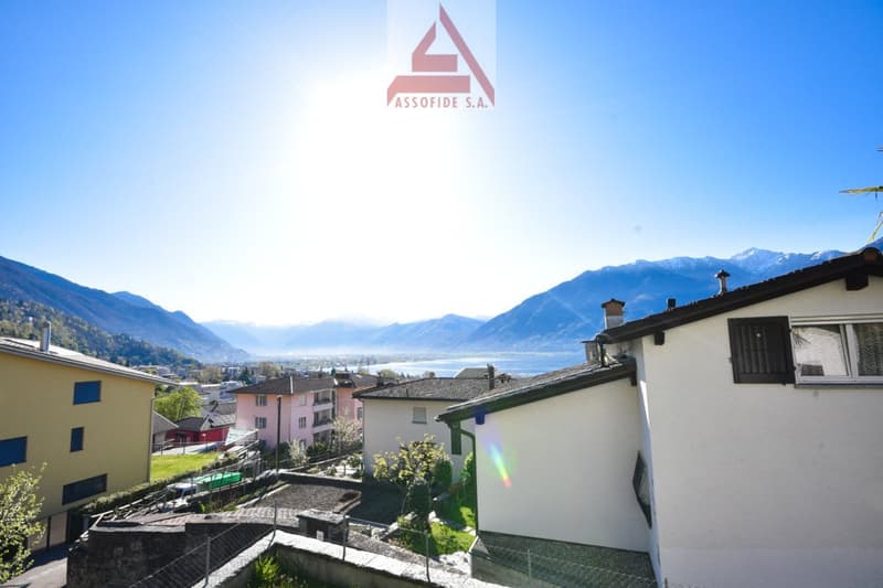 The image shows a view of a town or village nestled in the mountains, with a lake or body of water in the background. The buildings appear to be residential, with a mix of colors and architectural styles. The Assofide S.A. logo is visible at the top of th