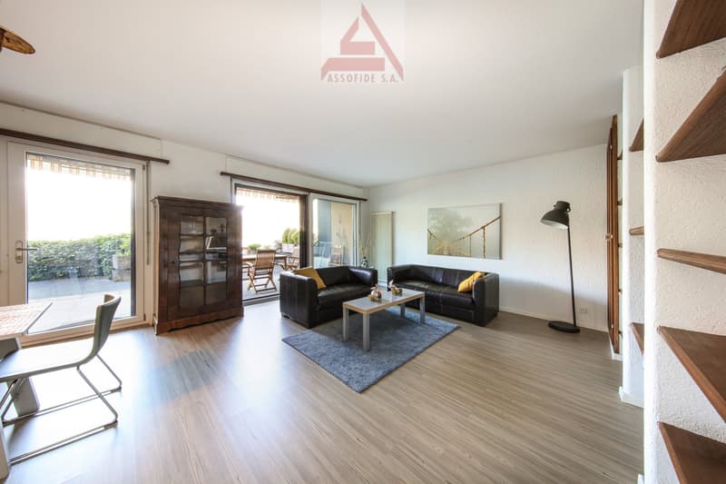 Spacious living room with hardwood flooring, large windows, a wooden cabinet, two leather sofas, a coffee table, and a floor lamp. The room has access to a balcony or terrace.