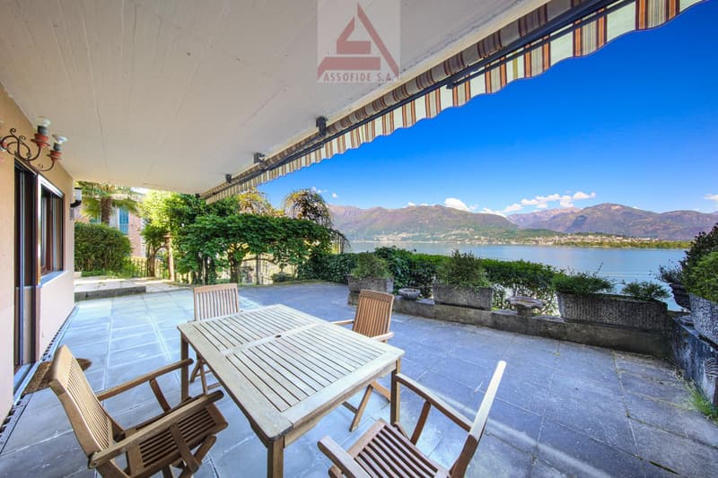 Outdoor patio with wooden table and chairs, overlooking a scenic lake and mountains in the background. The patio has a covered awning and is surrounded by lush greenery.