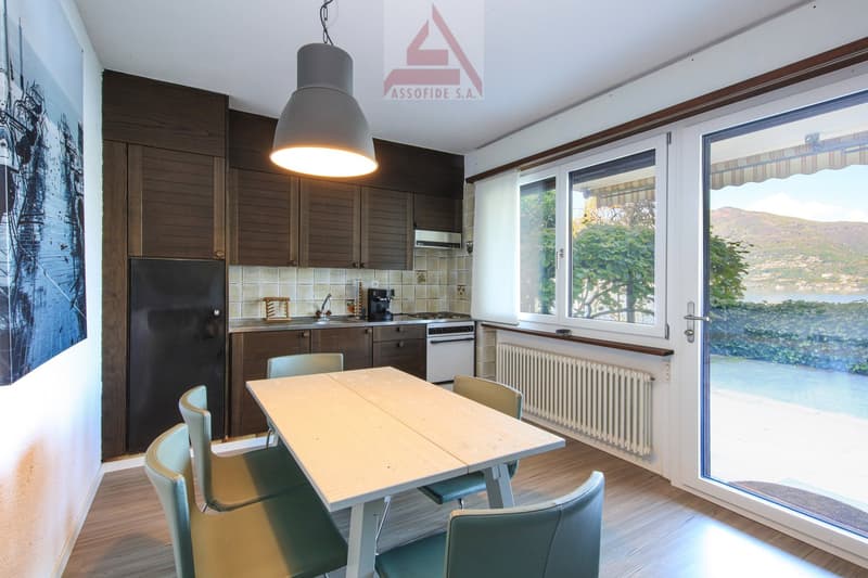 The image shows a modern kitchen and dining area with large windows providing a scenic view. The kitchen features dark wood cabinets, a tiled backsplash, and stainless steel appliances. The dining table is made of light-colored wood and is surrounded by m
