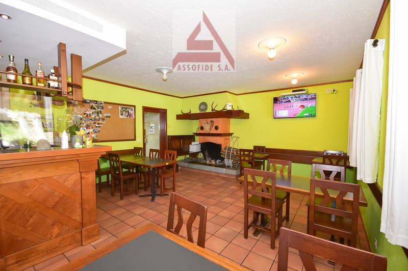 The image shows the interior of a restaurant or bar. The space has yellow walls, wooden furniture including tables and chairs, and a fireplace. There is a bar area with shelves displaying various alcoholic beverages. The room also has a TV mounted on the