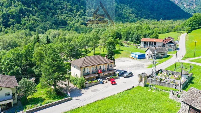 The image shows a small village nestled in a lush, green mountainous landscape. There are several buildings, including a red-roofed structure that appears to be a hotel or inn, as well as other residential and commercial buildings. There is a paved road r