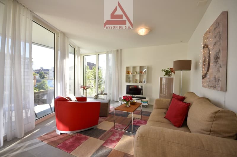 Spacious living room with large windows, red sofa, beige couch, wooden coffee table, shelving unit, and artwork on the wall. The room has access to a balcony or terrace.
