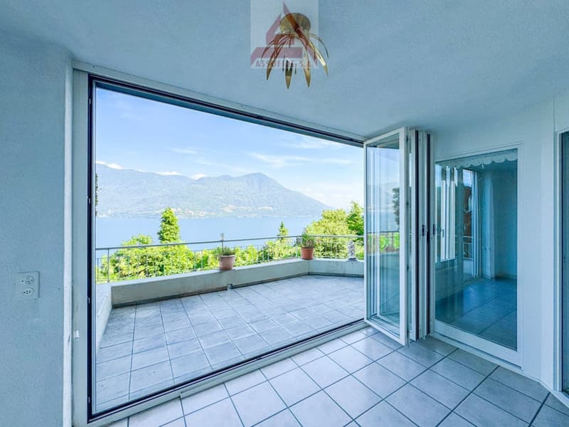 Spacious apartment with large balcony, offering a stunning view of the mountains and lake. The balcony has a tiled floor and is furnished with potted plants. The interior features large windows that provide abundant natural light and access to the balcony