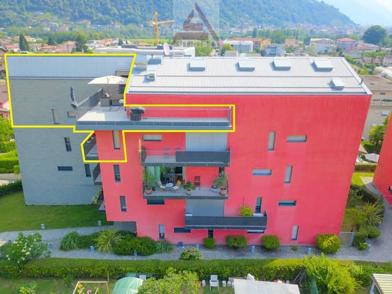 A modern, multi-story apartment building with a red exterior and balconies. The building has a flat roof and appears to be surrounded by greenery and landscaping.