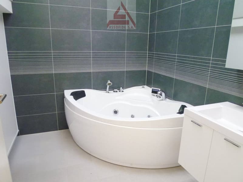 Large white corner bathtub with jets, grey tiled walls with horizontal striped pattern, white vanity cabinet