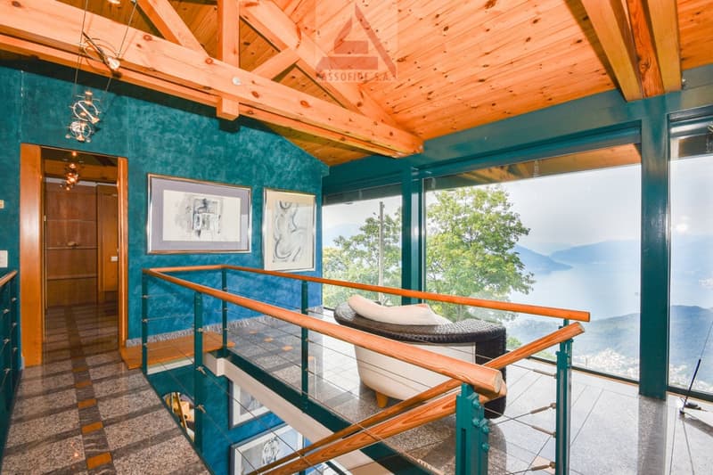 Spacious living room with wooden ceiling beams, teal walls, large windows offering a scenic view, and a cozy lounge area with a rattan chaise.