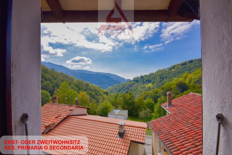 View of a mountainous, forested landscape from a window or balcony. The image shows red-tiled roofs of houses in the foreground, with a backdrop of lush green hills and mountains under a blue sky with clouds.