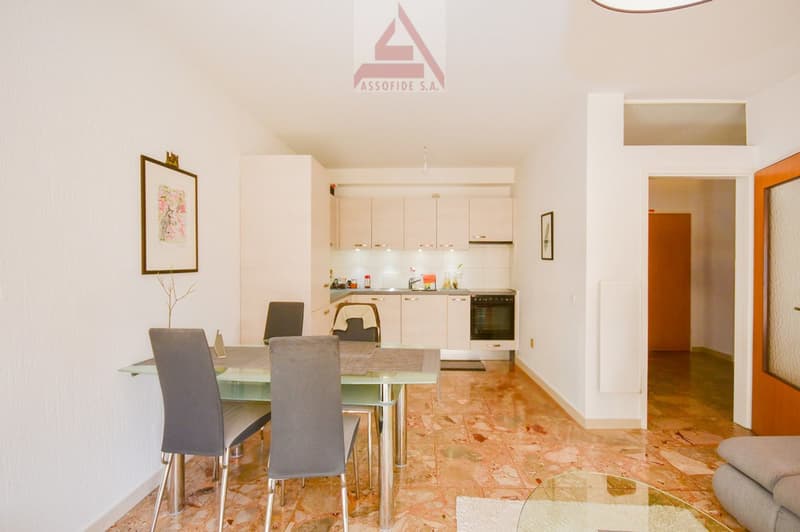 The apartment has an open-plan dining and living area with a modern kitchen, wooden floor, a dining table with four chairs, a glass top, and a glass side table. There is a picture frame on the wall, a radiator, and an entrance to another room. The kitchen
