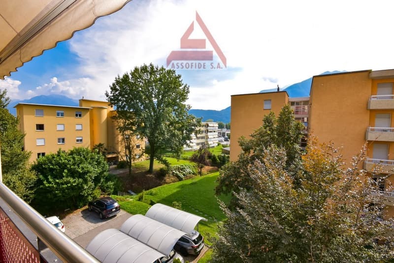 Outdoor area with parking shelter, trees, and a garden, overlooking the buildings and mountains in the distance.
