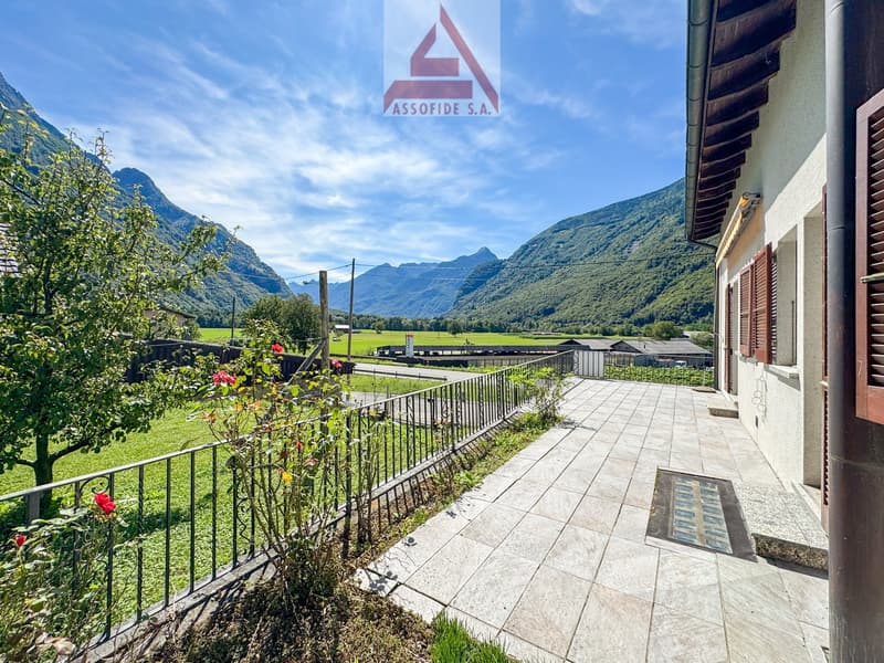 Exterior view of a house with a tiled terrace with plants and a view of mountains and fields.