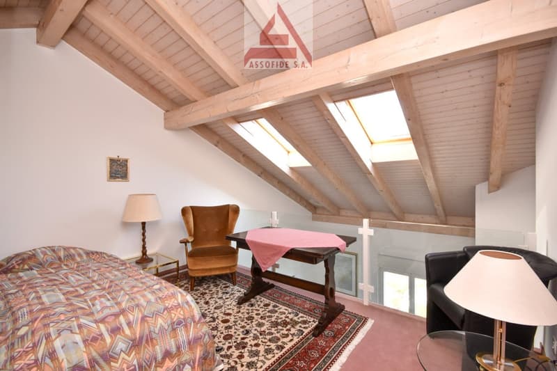 The bedroom features a bed, a wooden desk, a wooden chair, a lamp, and a couch. The ceiling has wooden beams and three skylights. There is a glass wall providing a view outside. A carpet is spread on the floor.