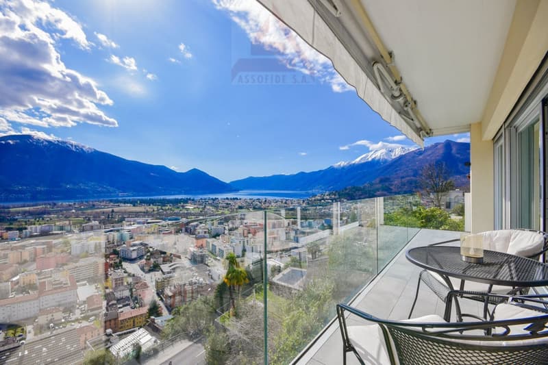 balcony with table and chairs, glass railing, mountain view, city view, clear blue sky, white clouds
