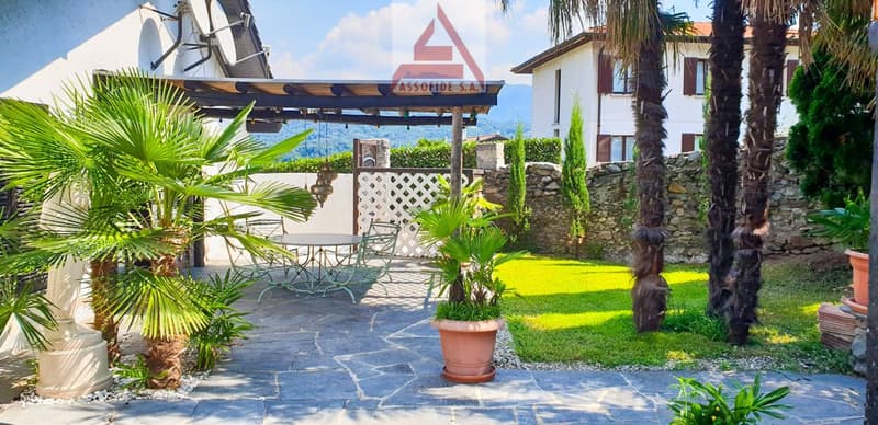 Outdoor patio with a white fence, palm trees, a round table, chairs, and a pergola. The backdrop features a stone wall, a mountain view, and a house.
