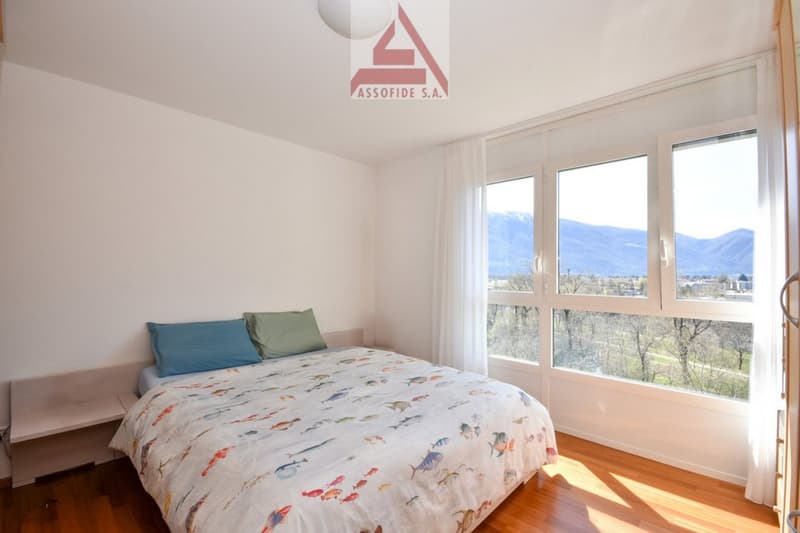 Bedroom with bed, pillows, nightstand, large windows, wooden floor, white walls, view of mountains and trees