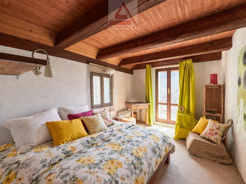 Bedroom with wooden ceiling, bed with floral cover, pillows, desk, chair, windows with yellow curtains, wicker chair, wooden cabinet, painting