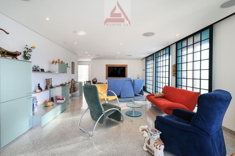 modern living room with white walls, gray floor, large windows, various chairs and a sofa, a TV, and decorative items on shelves