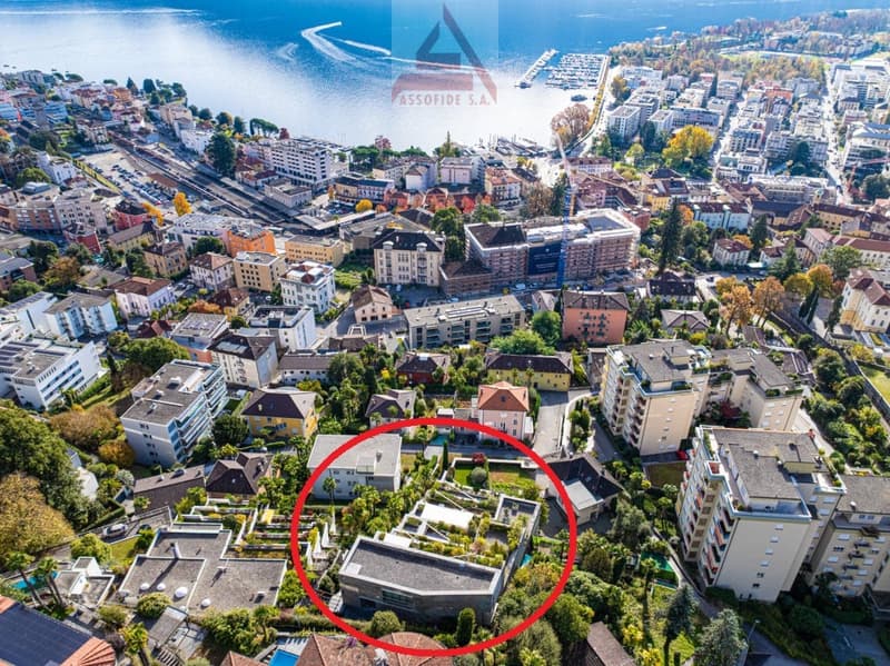 Aerial view of a city with many buildings, a red circle highlighting a specific building, a waterfront, and a lake in the distance.