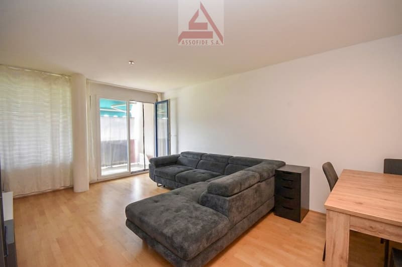 Living room with a gray sectional sofa, wooden floor, white walls, glass doors leading to a balcony, a wooden table, and a black cabinet.