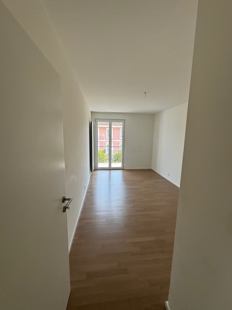 Empty room, wooden floor, white walls, glass door leading to balcony, power outlet on the wall.