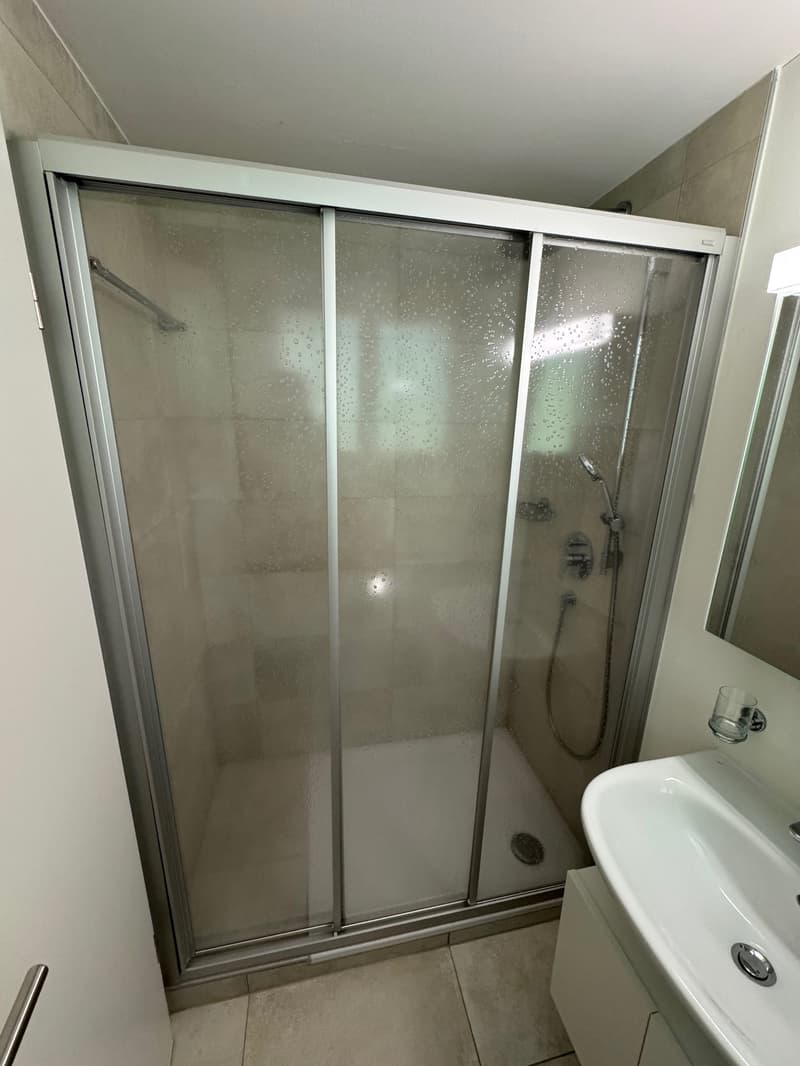 A bathroom with a glass shower enclosure, a sink, a mirror, and tiled floors.