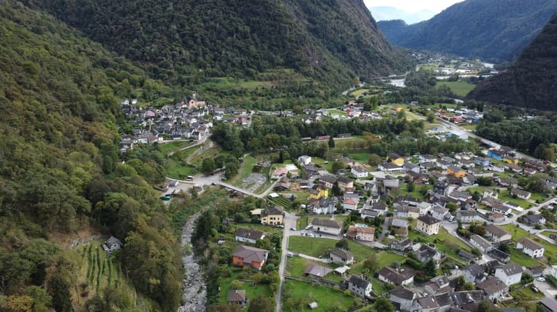 The image shows a picturesque mountain valley with a small town nestled among the lush green hills and forests. The town is surrounded by steep, forested slopes on both sides, with a river or stream running through the center. The buildings in the town ap
