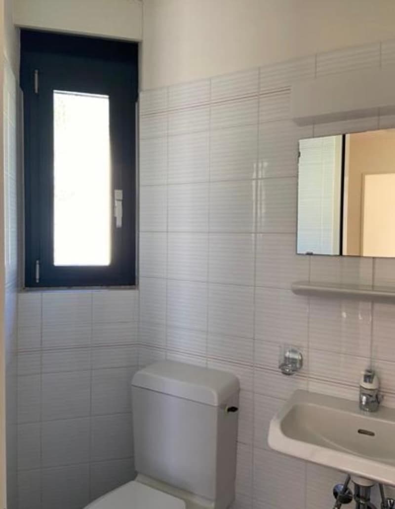 Small bathroom with white walls, white tiled floor, white sink, white toilet, mirror, and window