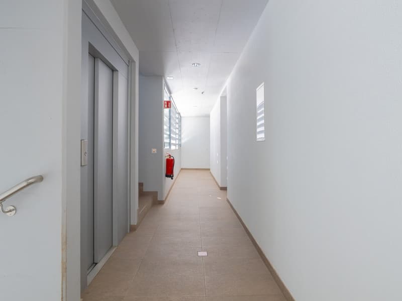 The image shows a long, narrow hallway with white walls and a tiled floor. There are sliding doors on the left side and a fire extinguisher mounted on the wall. The hallway appears to be well-lit with recessed lighting in the ceiling.