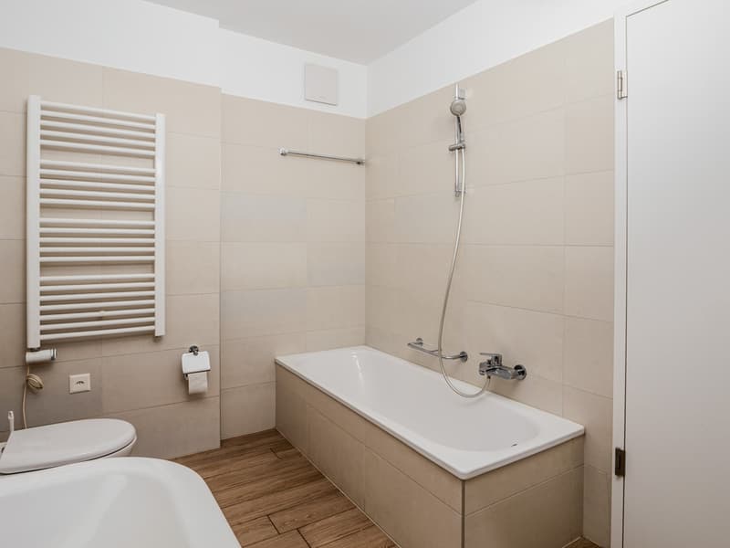 The image shows a modern bathroom with a white bathtub, a shower with a glass door, a white towel warmer, and beige tile walls.