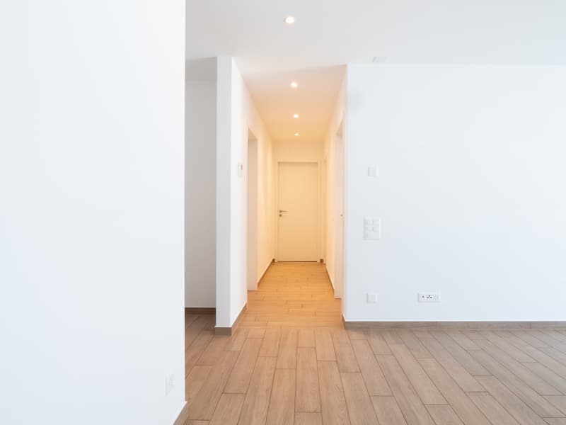 The image shows a long, well-lit hallway with white walls and a wooden floor. The hallway leads to a door at the end, and there are recessed ceiling lights illuminating the space.