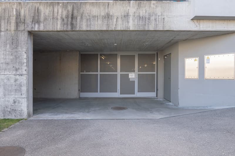 The image shows the exterior of a concrete building with a covered entrance. The entrance has large glass windows and a metal door. There is a concrete floor and a grassy area in front of the building.