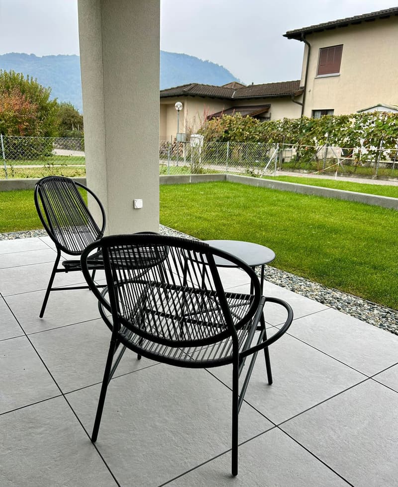 outdoor seating with 2 chairs, round table, tiled floor, green lawn, house and mountains in background