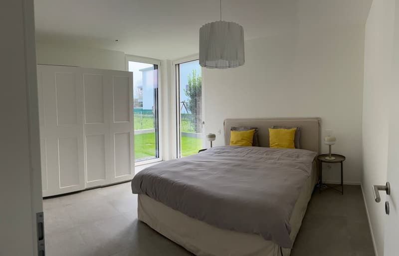 double bed, gray bedspread, two yellow pillows, sliding glass doors, view of grassy area, white ceiling lamp, white walls, white doors, wooden floor