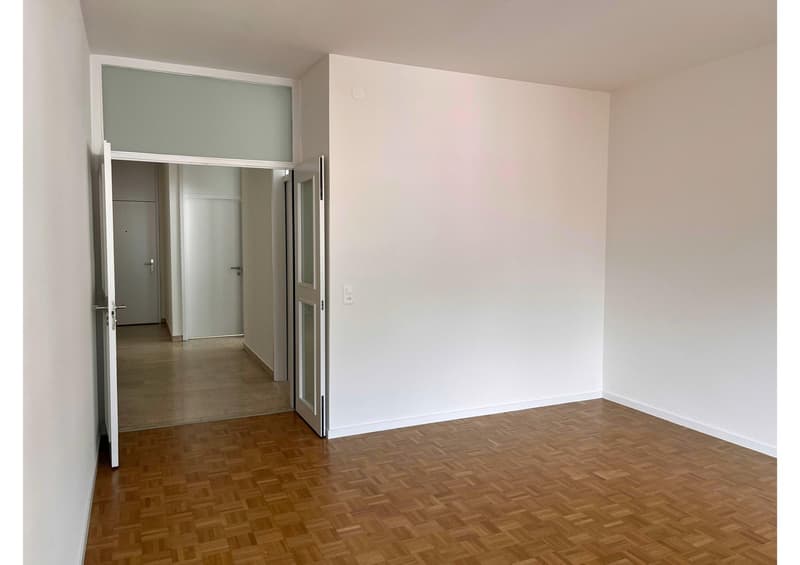Empty room with wooden floor, white walls, open door, and hallway leading to other rooms