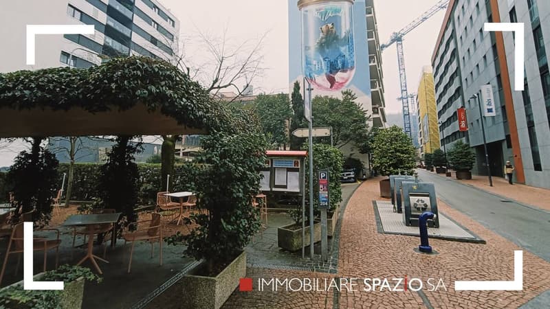 Outdoor cafe area with tables and chairs, shrubs, street with parked cars, high rise buildings, trees, large outdoor advertising banner, paved street