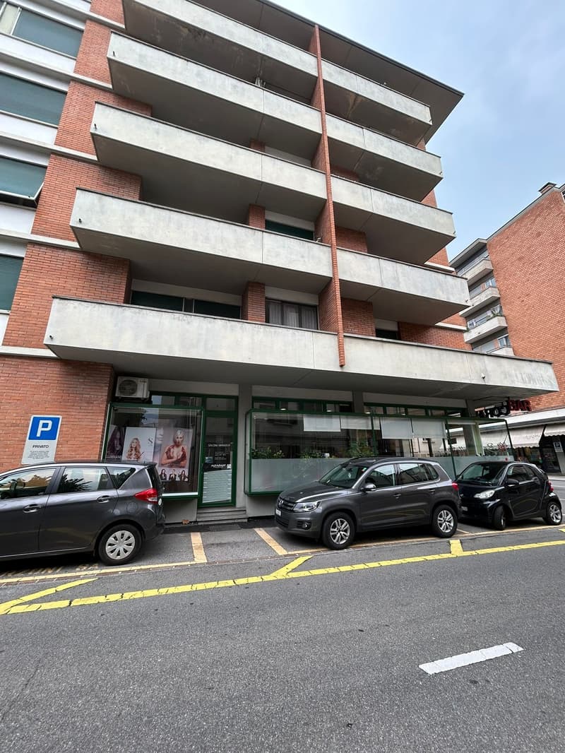 Multi-story brick and concrete building, multiple balconies, air conditioner on second floor, storefront with large windows, parking space, cars parked in front