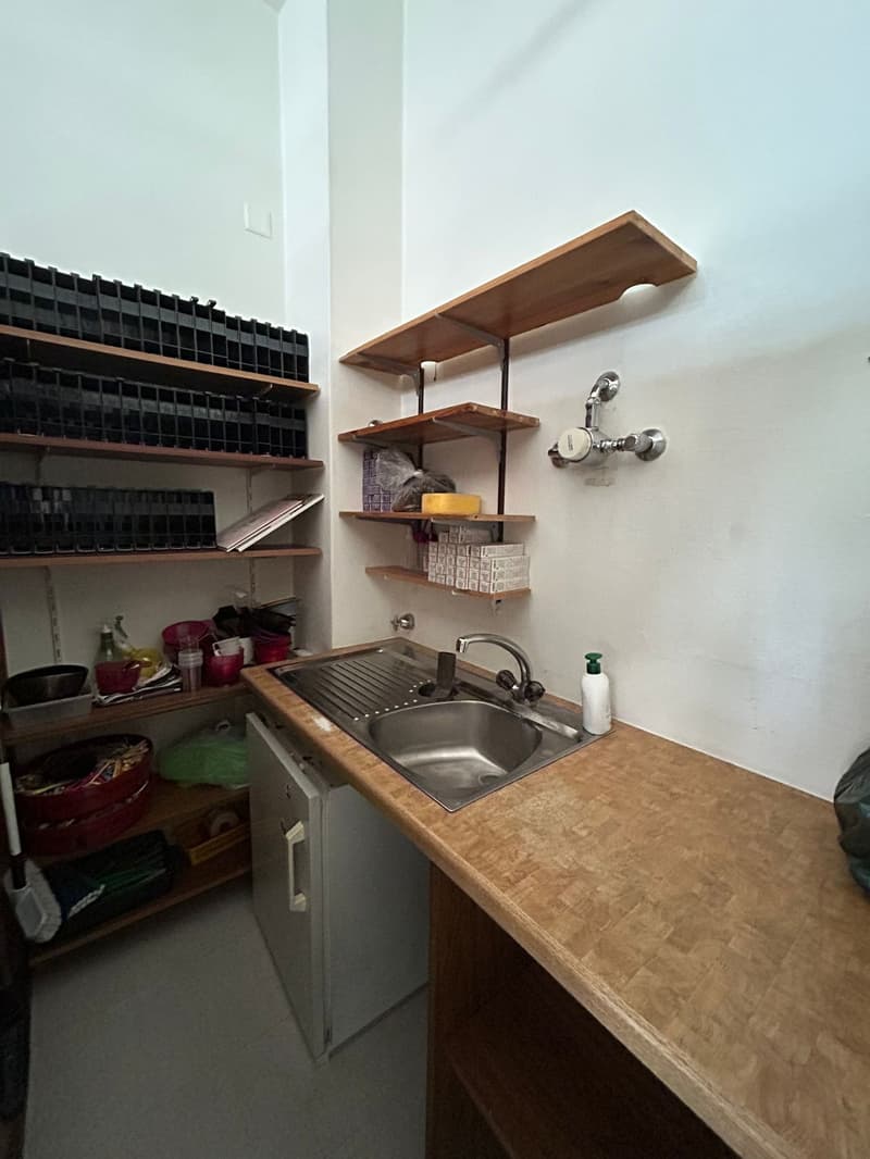 A small kitchen with wooden countertop, metal sink, white tiled floor, shelves, and a refrigerator.