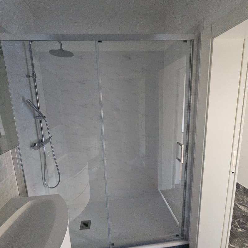 The image shows a modern bathroom with a glass-enclosed shower area, a minimalist design with gray tiled walls, and a shower head mounted on the wall.