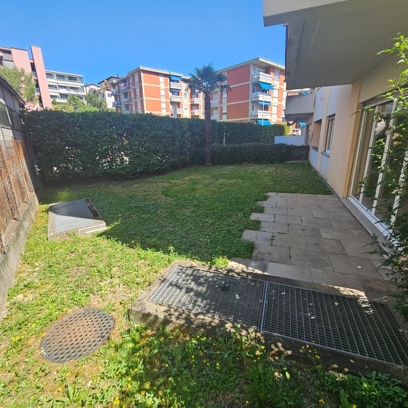 The image shows a residential apartment complex with several multi-story buildings. The buildings have a mix of orange and gray colors. There is a small grassy area with a palm tree in the foreground, and a paved walkway leading to the buildings. The comp
