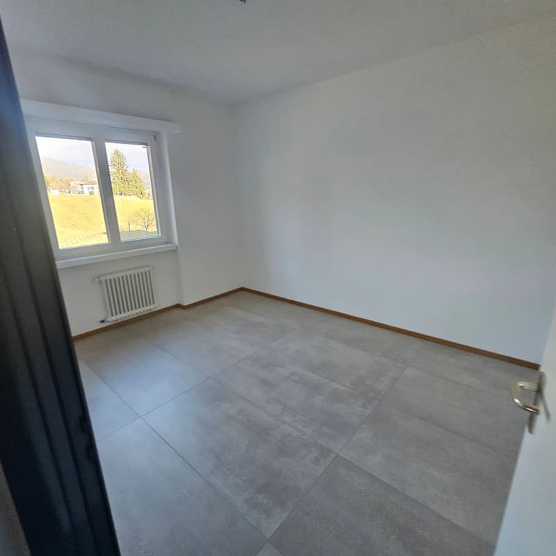 Empty room with white walls, gray tiled floor, large window with white frames, radiator.