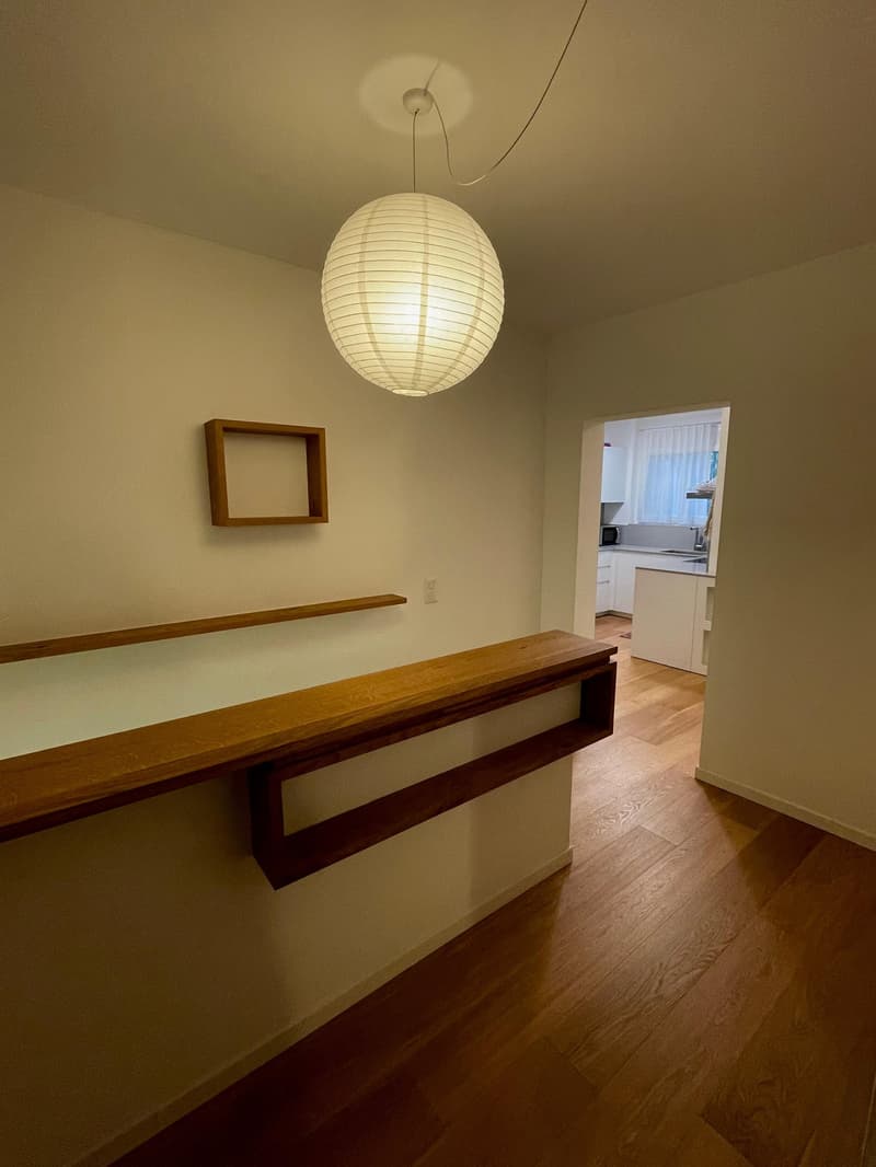 A room with wooden flooring and white walls, a large wooden table, a floating shelf, and a round paper lamp hanging from the ceiling. The room has an entrance to the kitchen with a microwave and sink.