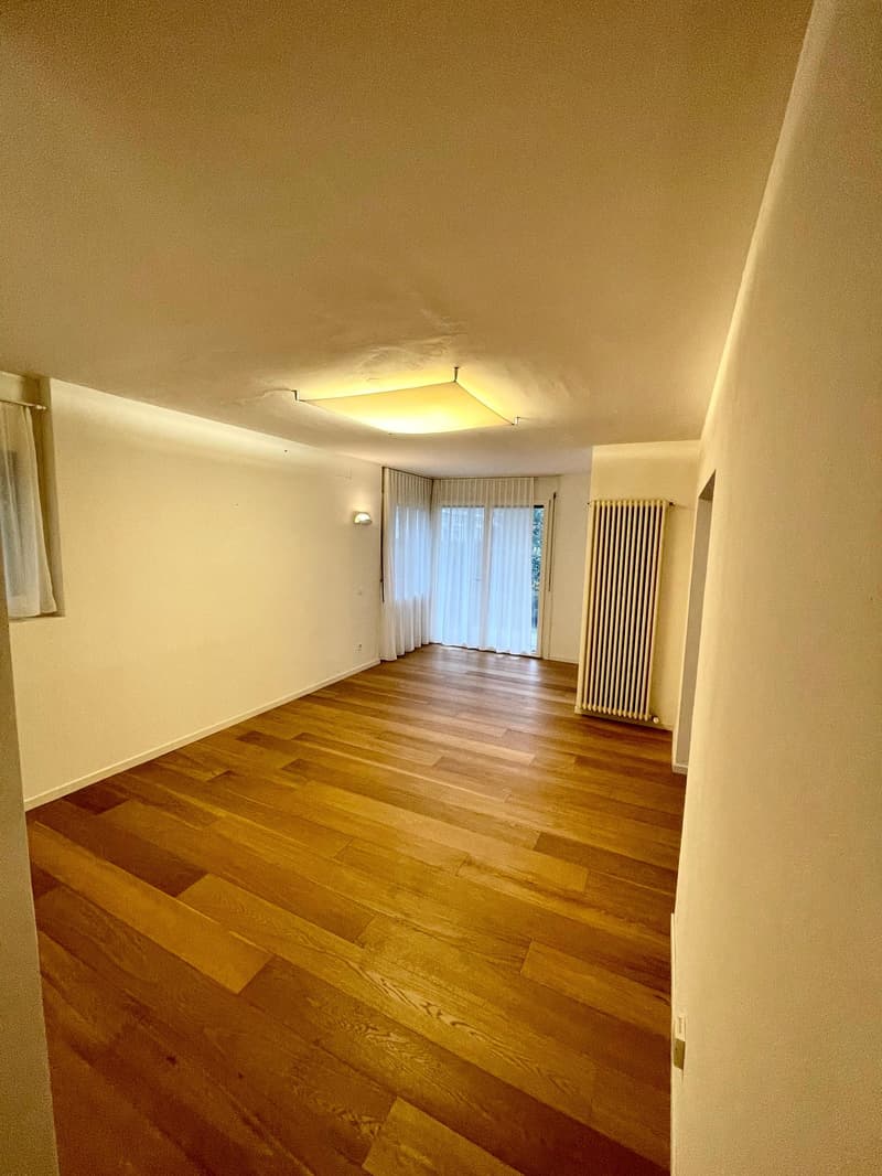 Empty room with wooden floor, white walls, ceiling lamp, and white curtains on the window.