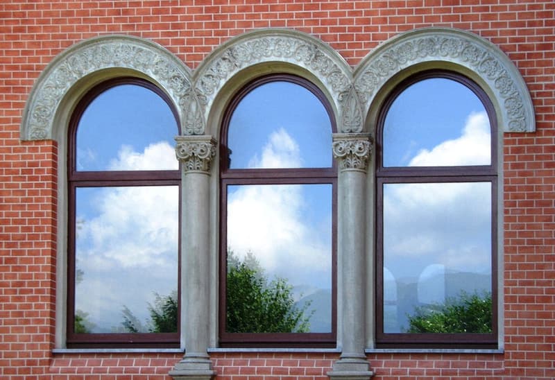 The image shows a brick building with three large arched windows. The windows have ornate stone frames and columns, and they reflect the cloudy sky and surrounding greenery outside. The building has a classic architectural style with a brick facade.