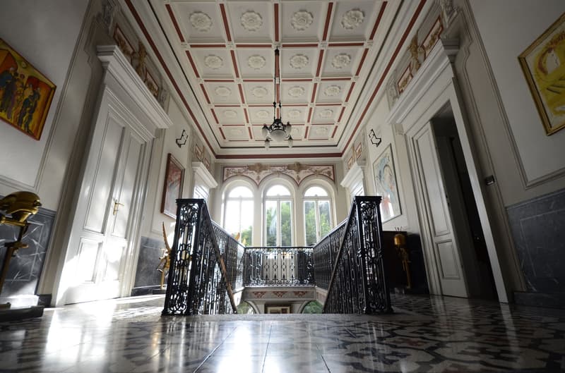Ornate and grand interior with intricate ceiling design, arched windows, and ornate iron railings on the staircase. The room has a formal and historic feel with the detailed architectural elements and decor.