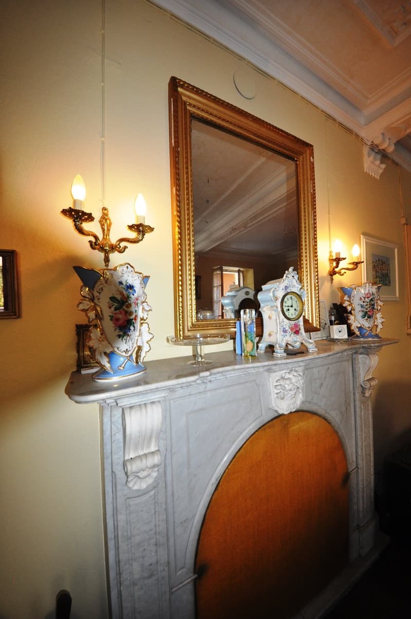 Ornate marble fireplace mantel with decorative vases and clocks, gilded framed mirror, wall sconces, and other antique decor items
