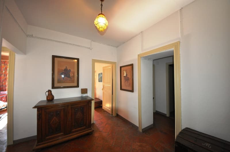 The image shows a hallway with white walls, a wooden cabinet with decorative elements, and framed artwork on the walls. The floor is covered with terracotta tiles. A decorative pendant light fixture hangs from the ceiling, providing warm lighting to the s