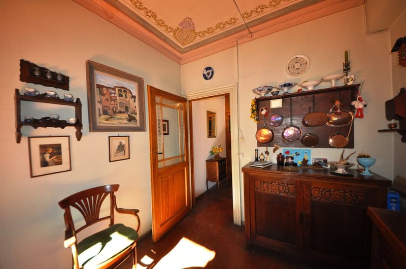 Ornate ceiling with decorative patterns, wooden furniture including a cabinet with decorative elements, framed artwork on the walls, and a fireplace visible in the room.